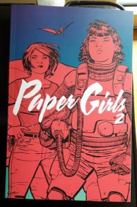 Zwei der Paper Girls in pink vor blauem Grund und einem Flugsaurier im Hintergrund