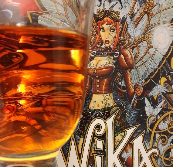 Weinglas mit orangefarbener Flüssigkeit im Vordergrund vor Cover des Comics Wika Band 2, das eine Fee zeigt