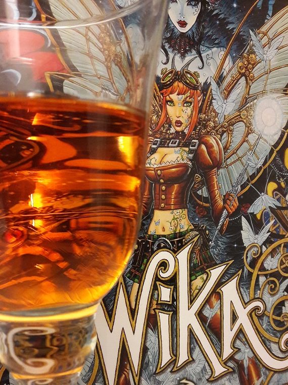 Weinglas mit orangefarbener Flüssigkeit im Vordergrund vor Cover des Comics Wika Band 2, das eine Fee zeigt