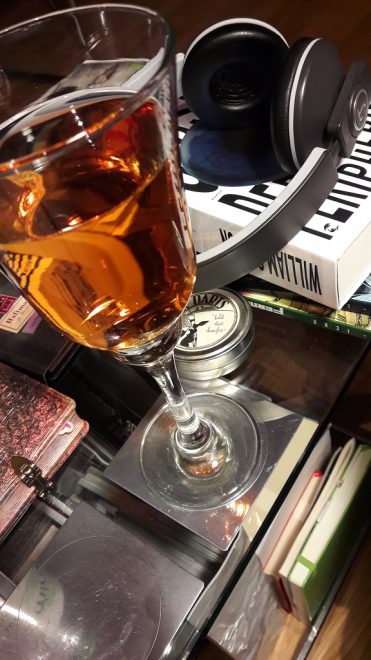 Glas mit orangefarbener Flüssigkeit im Vordergrund, dahinter das Buch und darauf ein Kopfhörer