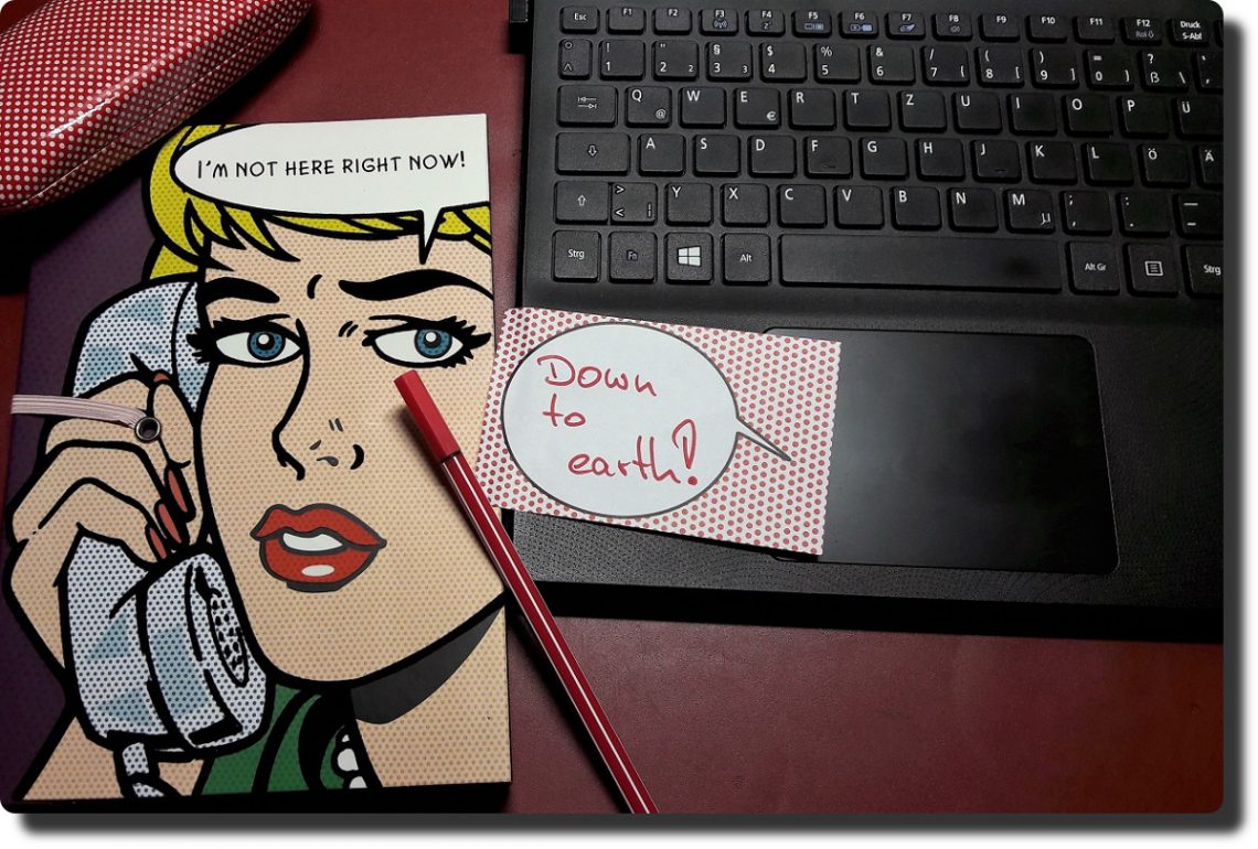 Frau in Comic-Style auf einem Notizbuch, ein roter Stift, ein Klebezettel mit Aufschrift "Down to earth!" und ein Notebook
