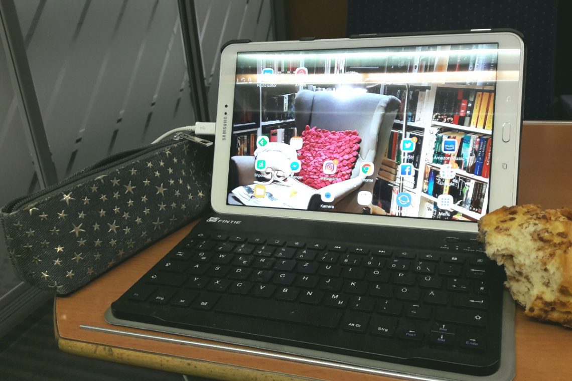 Tablet mit Tastatur, daneben eine Faulenzermappe und ein Brötchen im Zug