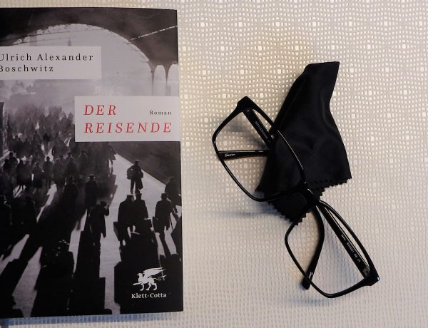 Buch liegt neben einer schwarzen Brille und einem Tuch auf einem hellen Untergrund