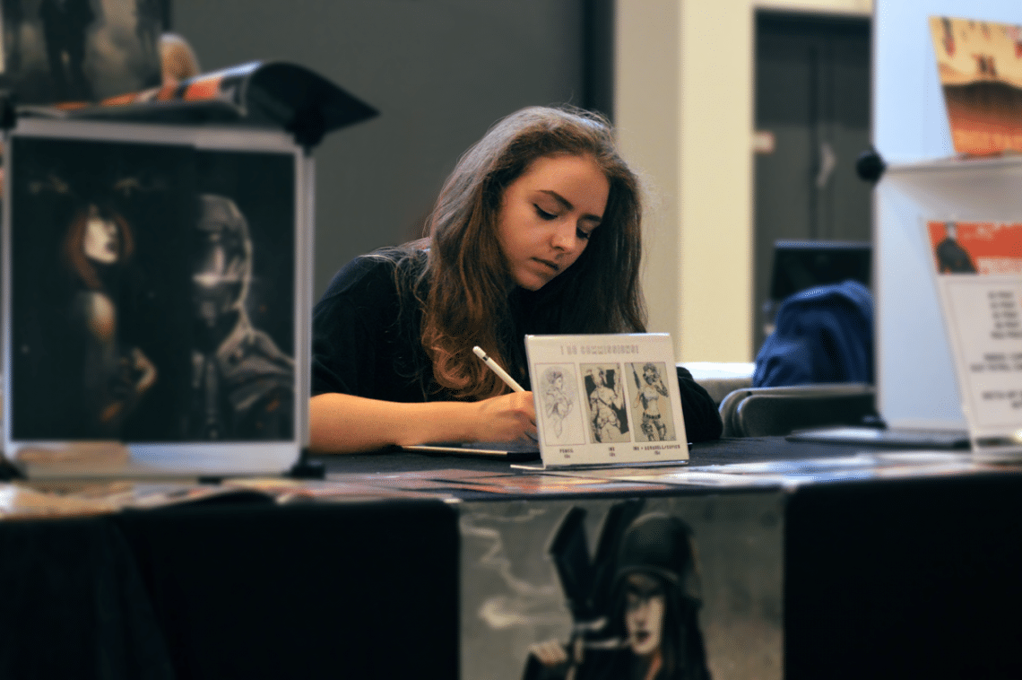 Junge Frau sitzt an einem Tisch inmitten ihrer Zeichnungen und arbeitet konzentriert
