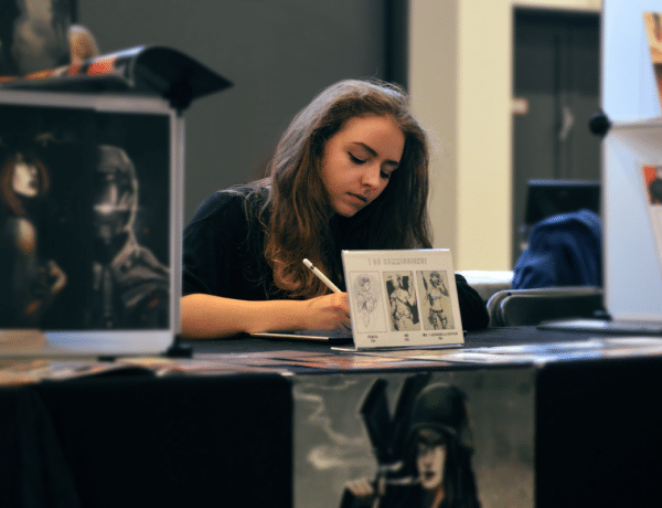 Junge Frau sitzt an einem Tisch inmitten ihrer Zeichnungen und arbeitet konzentriert