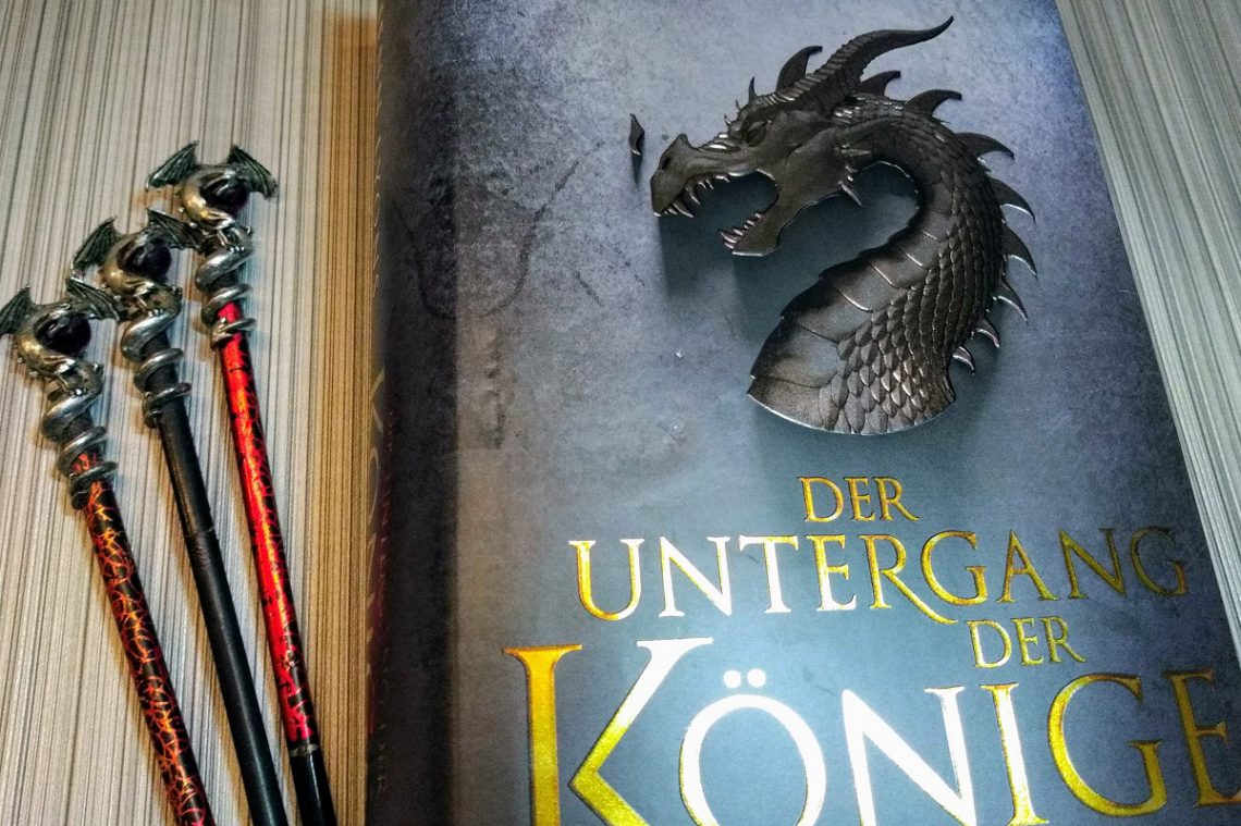 Buch neben drei Stäben an denen oben silberne Drachen befestigt sind