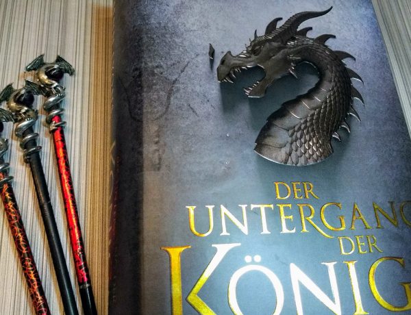 Buch neben drei Stäben an denen oben silberne Drachen befestigt sind