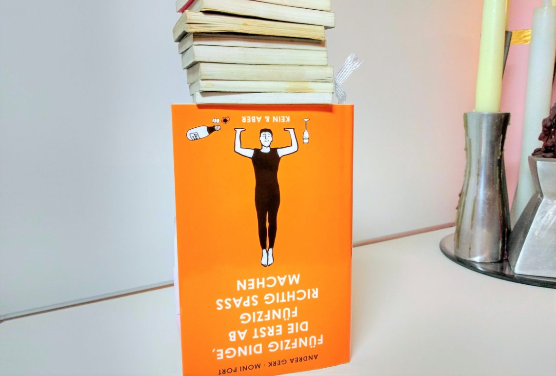 Orangefarbenes Buch steht auf dem Kopf und obendrauf liegen kleine Minibücher mit dem Buchschnitt davor. Auf dem Cover eine Frau, die diese kleinen Bücher zu tragen scheint