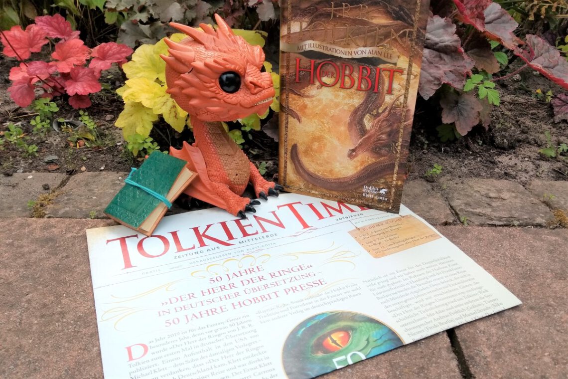 Funko Figur von Smaug sitzt auf einer Ausgabe der Zeitung Tolkien Times, daneben die illustrierte Fassung vom Hobbit mit einem feuerspeienden Drachen darauf - das alles vor Pflanzen