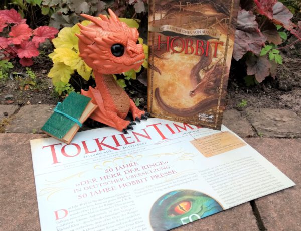 Funko Figur von Smaug sitzt auf einer Ausgabe der Zeitung Tolkien Times, daneben die illustrierte Fassung vom Hobbit mit einem feuerspeienden Drachen darauf - das alles vor Pflanzen