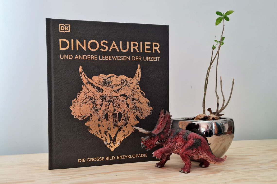 Buch Dinosaurier steht neben einer Dinosaurierfigur und daneben eine Pflanze in silbernem Topf