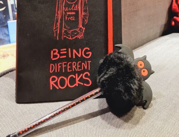 Schwarzes Notizbuch mit Aufschrift Being different rocks, davor ein schwarzroter Kugelschreiber, auf dem oben eine schwarze Plüschfledermaus steckt