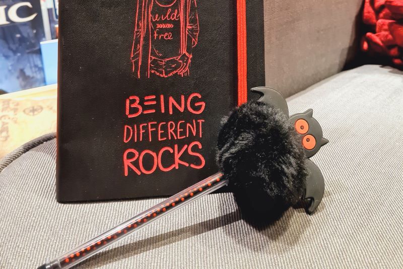 Ich bin jetzt bei VG Wort registriert Schwarzes Notizbuch mit Aufschrift Being different rocks, davor ein schwarzroter Kugelschreiber, auf dem oben eine schwarze Plüschfledermaus steckt