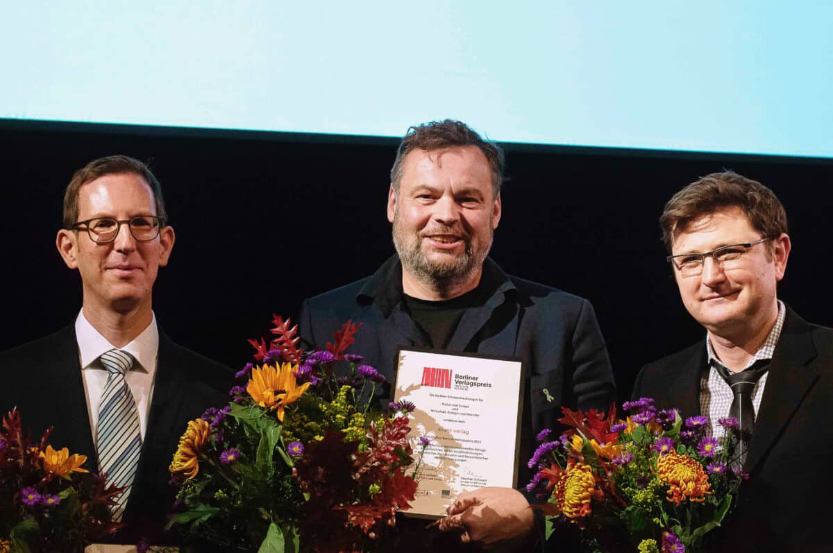 avant verlag erhält den berliner verlagspreisDrei Männer nebeneinander, die frontal in die Kamera schauen. Mann mit kurzem Haar in der Mitte hält eine Urkunde und einen Blumenstrauß in der Hand