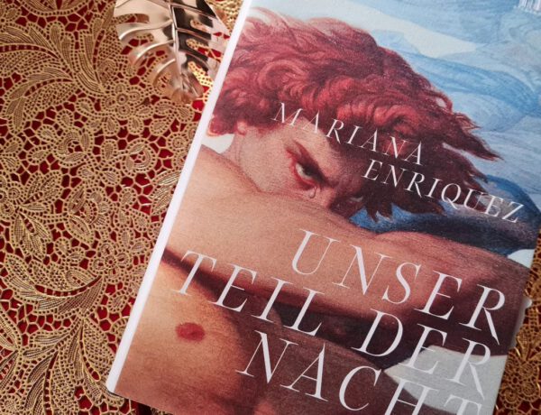 Roman Unser Teil der Nacht von Mariana Enriquez liegt auf einem goldenen Untergrund mit Spitzenoptik. Auf dem Cover ein rothaariger Mann mit freiem Oberkörper, der einen Arm vor ein Gesicht hält