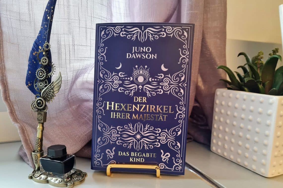 Buch mit Aufschrift Der Hexenzirkel Ihrer Majestät steht neben einer Schreibfeder und einem Tintenglas und einem Blumetopf mit Grünpflanze