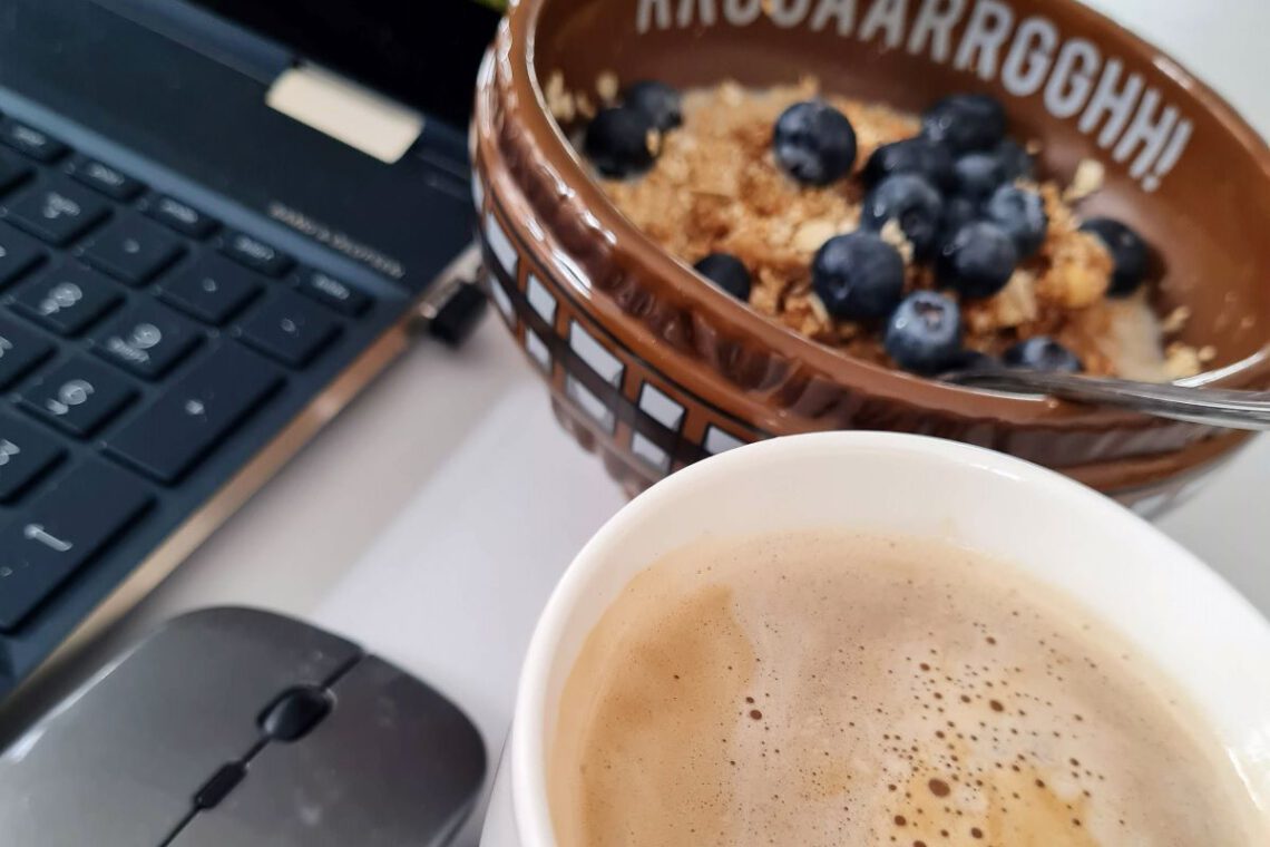 Kaffeetasse unten rechts, darüber eine braune Müslischale mit Heidelbeeren und links sind eine Computermaus und ein Notebook im Ausschnitt zu sehen