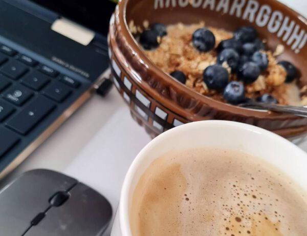 Kaffeetasse unten rechts, darüber eine braune Müslischale mit Heidelbeeren und links sind eine Computermaus und ein Notebook im Ausschnitt zu sehen