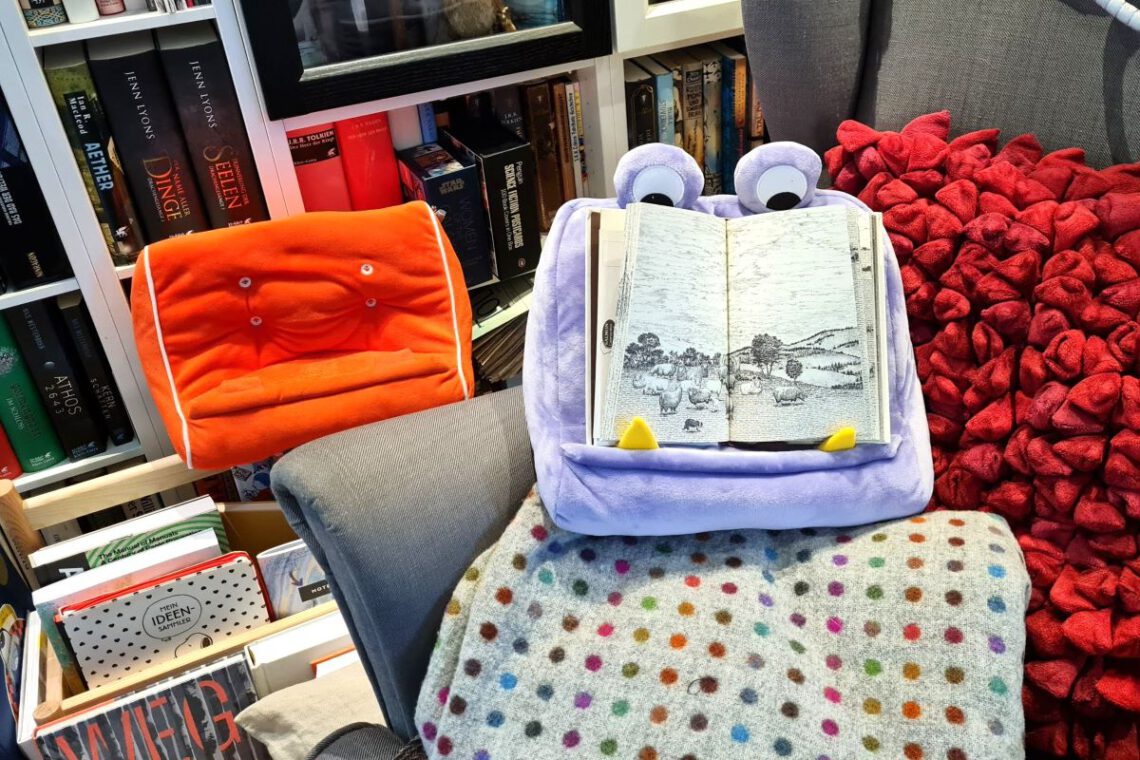 BookMonster Lesekissen in lila mit zwei Augen oben und darauf ein aufgeschlagenes Buch. Das Ganze auf einem Sessel mit Wolldecke vor einem Bücherregal BookCouch im Hintergrund