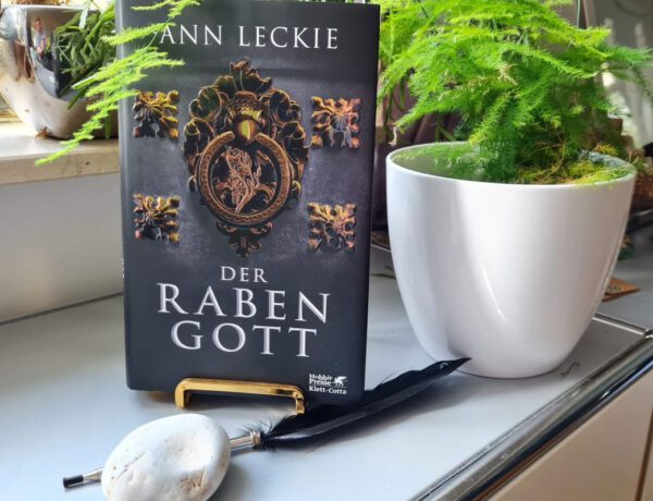 Buch steht auf einem goldenen Buchständer, davor eine schwarze Feder und ein Stein, im Hintergrund Pflanzen