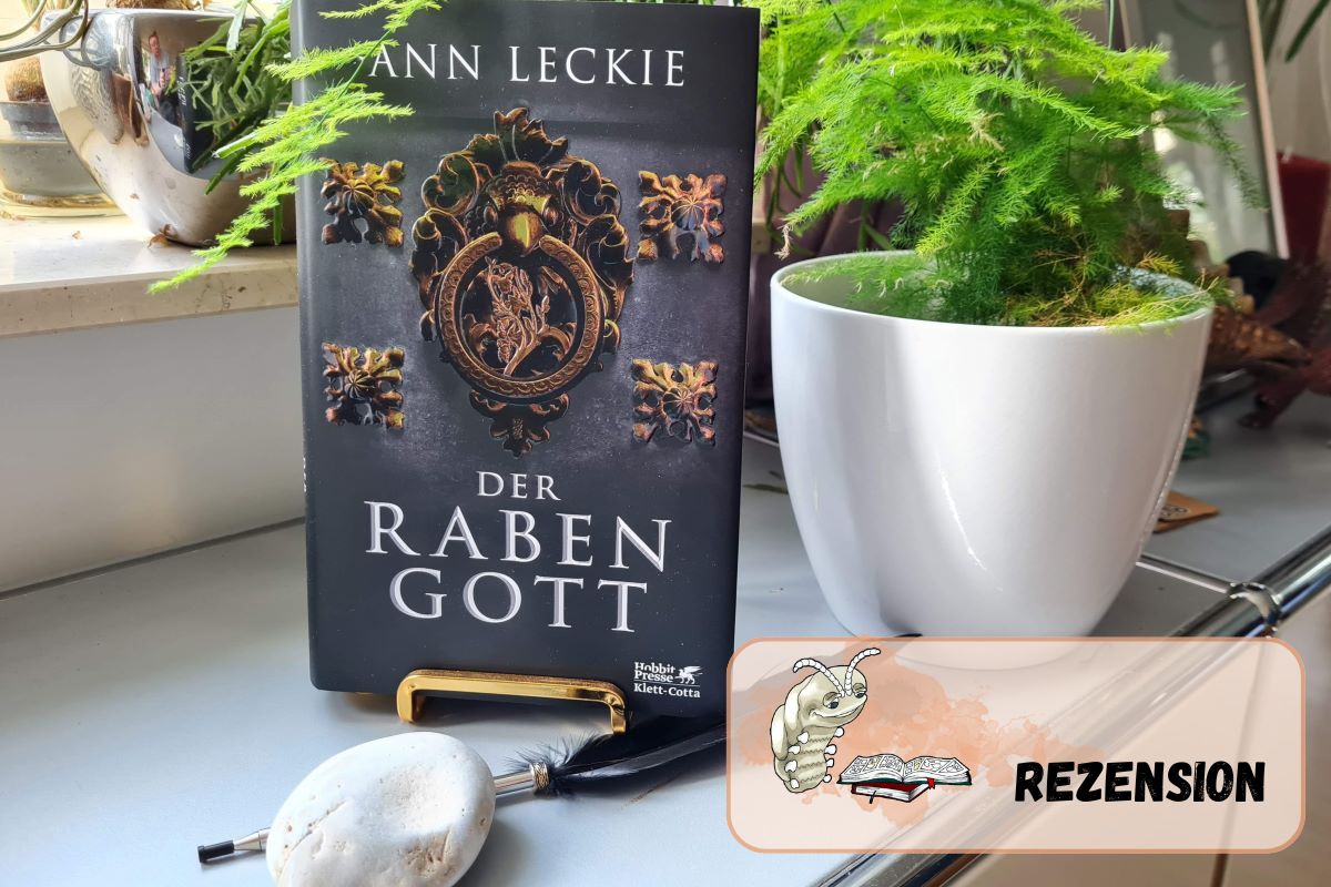 Buch steht auf einem goldenen Buchständer, davor eine schwarze Feder und ein Stein, im Hintergrund Pflanzen