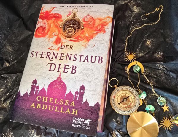 Sternenstaubdieb Chelsea Abdullah - Roman liegt auf einem schwarzen Papier, daneben goldener Glitter, ein Kompass und eine Kristallkette