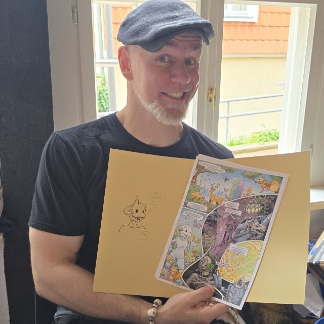 Mann hält Comicseiten in der Hand und lächelt, im Hintergrund ein Fenster