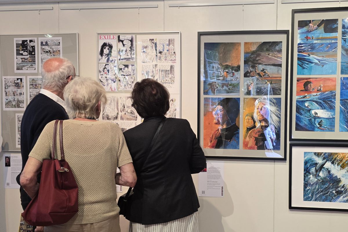 Drei Menschen schauen auf an der Wand hängende Comics im Rahmen einer Ausstellung